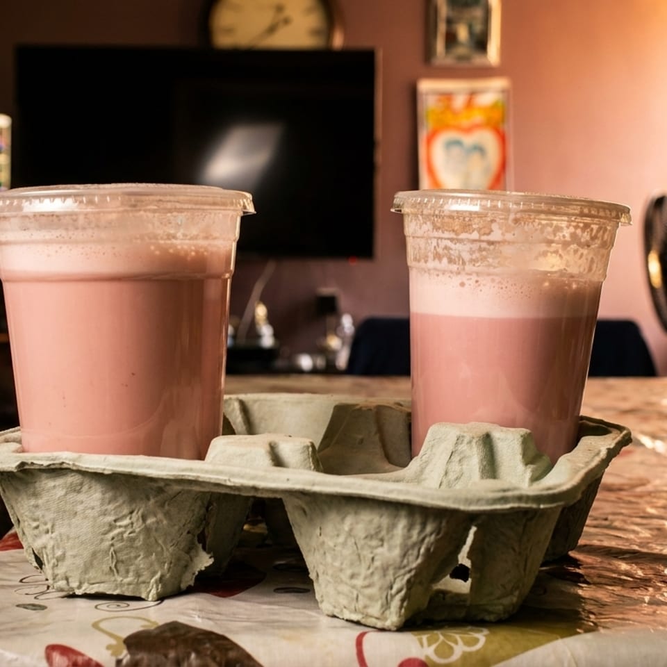 Two pink milkshakes in plastic cups, in a cardboard carrier, on a patterned table.
