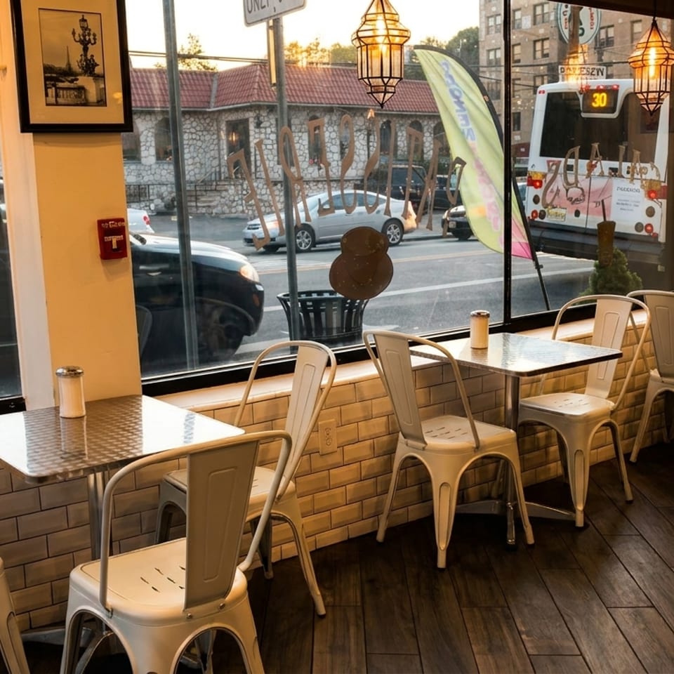 Cozy cafe interior with white chairs and metal tables by a large window overlooking a city street.